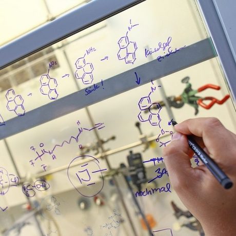 Eine Hand schreibt Formeln und chemische Verbindungen für ein Experiment an die Glasscheibe in einem Labor.