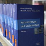 Seitliche Aufnahme eines Regals in einer Bibliothek, in dem blaue Bücher aufgereiht sind. Das vorderste Buch trägt den Titel "Kostenrechnung und Kostenanalyse".