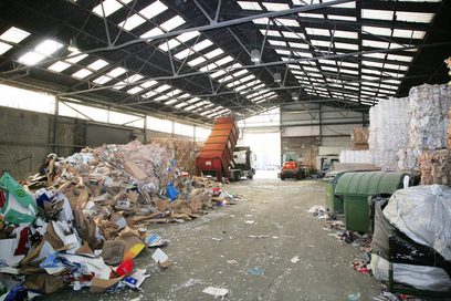 Blick vom Ende einer Recyclinghalle in Richtung Hallentor, dort lädt ein LKW Müll ab. In der Halle befinden sich auf der rechten Seite sortierte Wertstoffe, auf der linken Seite verschiedene Abfallberge.