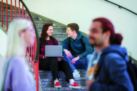 Zwei Studierende sitzen auf einer Treppenstufe nebeneinander und lächeln sich an.