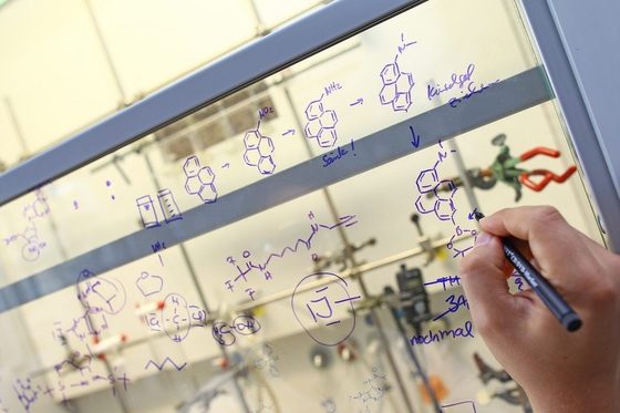Eine Hand schreibt Formeln und chemische Verbindungen für ein Experiment an die Glasscheibe in einem Labor.