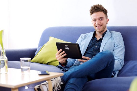 Junger Mann sitzt auf einer Couch und arbeitet an einem Tablet.