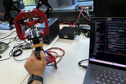Ein mechanischer Greifarm auf einem Schreibtisch, daneben steht ein Laptop mit einer Code-Oberfläche auf dem Bildschirm..