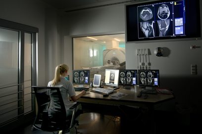 Eine junge Frau in weißem Kittel überwacht die Untersuchung von Erkrankten in einem Magnetresonanztomographen durch das Fenster eines nebengelegenen Kontrollraums.