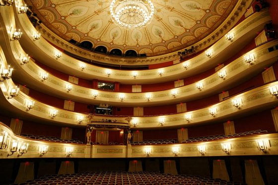 Blick von der Bühne in einen hell erleuchteten Opernsaal mit Logenplätzen über vier Etagen.