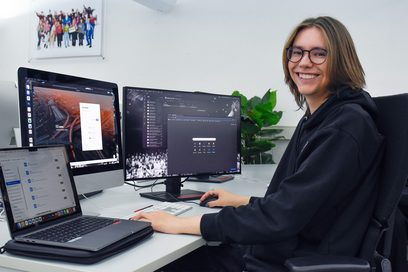 Ein junger Mann sitzt an einem Schreibtisch vor zwei Bildschirmen sowie einem aufgeklappten Laptop und bearbeitet digital einen Kundenauftrag.