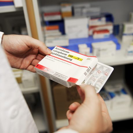 Eine Person im weißen Kittel hält eine Medikament-Schachtel vor einem Schrank mit Medikamenten.