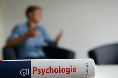 Ein Psychologie Lehrbuch ist im Vordergrund zu sehen, während eine Person verschwommen im Hintergrund zu sehen ist.