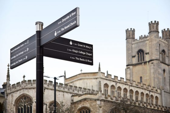Nachaufnahme eines Campus-Wegweisers an der Universität von Cambridge in England. Im Hintergrund ist ein Teil der des historischen Gebäudes der Universität mit einem Turm zu sehen. (Foto: Sonja Trabandt | Bundesagentur für Arbeit)