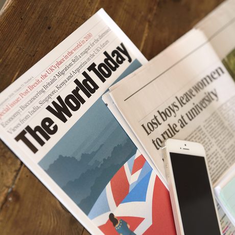 Auf einem Tisch liegt ein internationales Magazin, eine Zeitung mit englischer Überschrift und ein weißes iPhone.