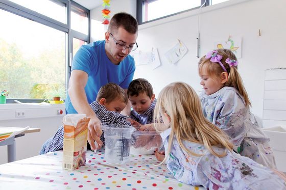 Ein junger Mann mit Brille, bekleidet mit einem blauen T-Shirt leitet zwei Mädchen und zwei Jungen, an einem Tisch mit bunt gepunkteter Tischdecke, bei der Herstellung eines Knetteiges an. Die Kinder haben die Hände in einer Mehlschüssel, der junge Mann greift an einen Messbecher mit Wasser. 