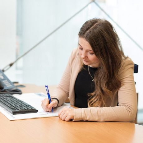 Eine Frau im Business-Outfit sitzt an einem Tisch und schreibt auf einen Block Papier.