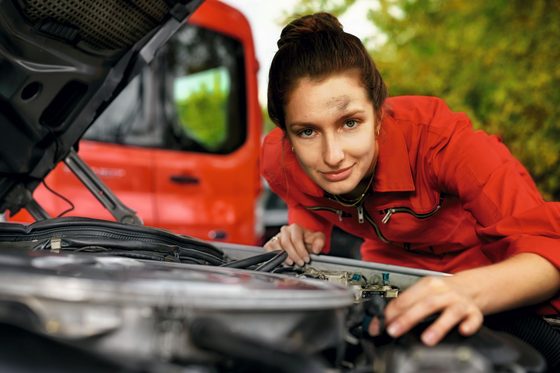 Eine junge Frau lehnt sich über die geöffnete Motorhaube eines Autos. 
