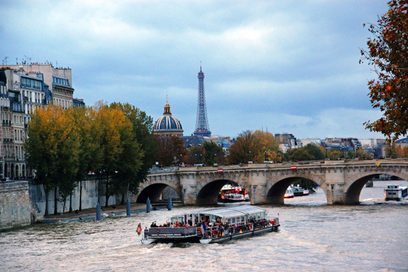 Panoramablick auf Paris, von der Seine aus fotografiert. Im Vordergrund ein Boot voller Passagiere in der Nähe einer alten Brücke. Zwischen den Bäumen mit herbstlichen Farben, steht der Invalidendom. Im Hintergrund der Eiffelturm. Der Himmel über Paris ist bewölkt.