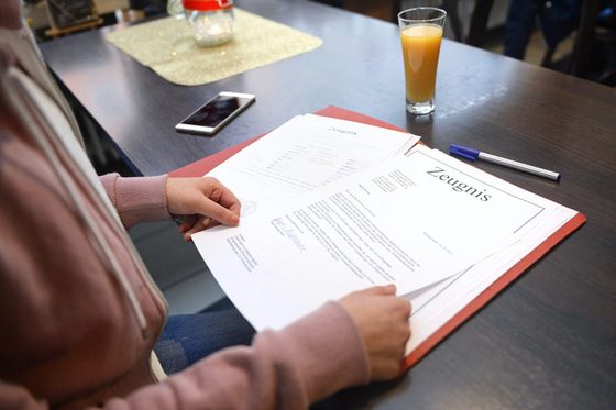 Frauenhände blättern in einer Mappe mit Dokumenten, auf denen "Zeugnis" steht. Die Mappe liegt auf einem großen Tische, Auf dem neben einem Kugelschreiber ein Glas Orangensaft steht. Ein Smartphone liegt ebenfalls daneben.