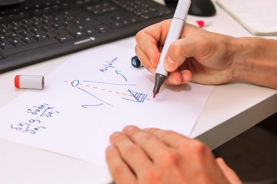 Auf einem Schreibtisch vor einer schwarzen Tastatur schreibt eine Männerhand mit Rotstift auf ein Dokument, auf dem bereits eine Formel und eine Grafik notiert sind.