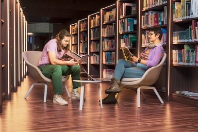 Eine Frau und ein Mann sitzen sich in einer Bibliothek gegenüber und lesen in Büchern.