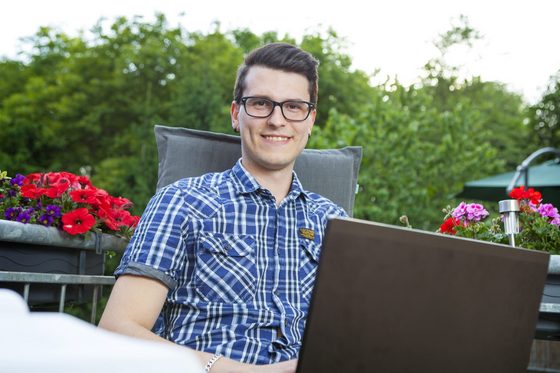 Ein junger Mann sitzt mit seinem Laptop auf einem Balkon.