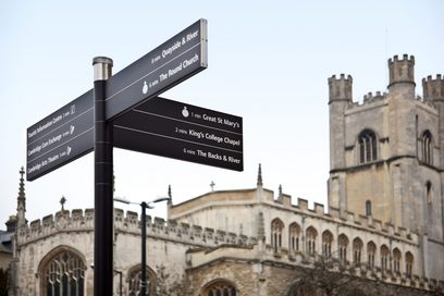 Nachaufnahme eines Campus-Wegweisers an der Universität von Cambridge in England. Im Hintergrund ist ein Teil der des historischen Gebäudes der Universität mit einem Turm zu sehen.