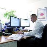 Ein junger Mann sitzt an einem Schreibtisch vor einem Computer und hat eine Mappe vor sich aufgeschlagen.