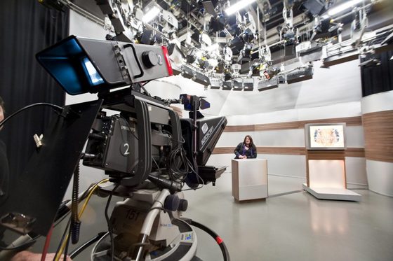 In einem Fernsehstudio, Nahaufnahme einer Fernsehkamera, die auf eine im Hintergrund stehende Frau gerichtet ist. Zahlreiche Strahler hängen von der Decke.