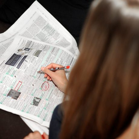 Das Foto zeigt eine junge Frau von hinten, die Wohnungsanzeigen in einer Tageszeitung mit Rotstift markiert.