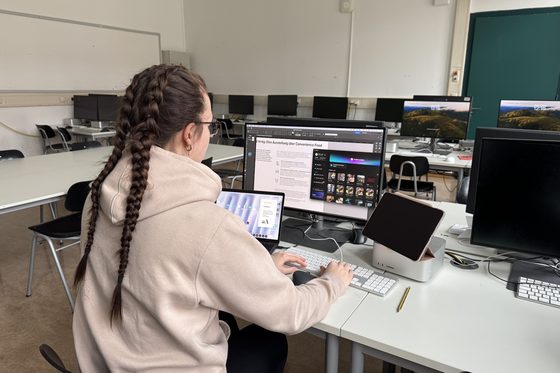 Rückansicht einer jungen Frau am Arbeitsplatz, die vor mehreren Bildschirmen sitzt und arbeitet. 