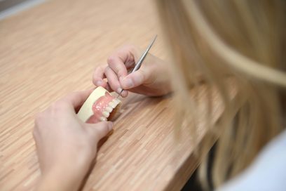	Eine Frau hält Skalpell mit sehr dünner Klinge ab ein künstliches Gebiss über einen Tisch.