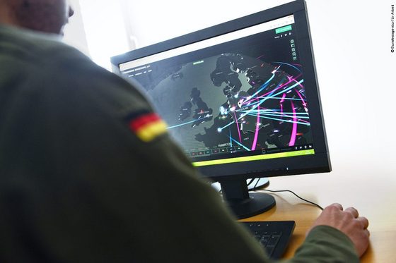 Ein Mann in Bundeswehruniform sitz vor einem Computerarbeitsplatz und schaut auf einen Monitor. In seiner rechten Hand hält er eine Computermaus. Auf dem Bildschirm ist eine virtuelle Weltkugel zu sehen, auf der rosa- und blaufarbige Linien laufen.