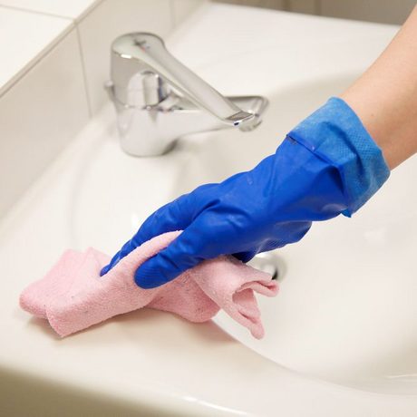 Jemand reinigt in einem blauen Handschuh ein Waschbecken mit einem rosa Lappen.