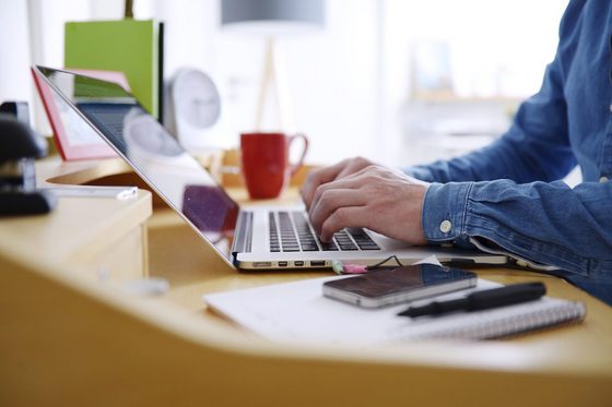 Nahaufnahme der Hände eines jungen Mannes in einem Jeanshemd, der auf einem hellgrauen Laptop tippt. Auf dem Schreibtisch liegen auch ein Notizblock, ein Handy und ein Stift sowie im Hintergrund eine Uhr, eine Tischlampe und eine Teetasse.