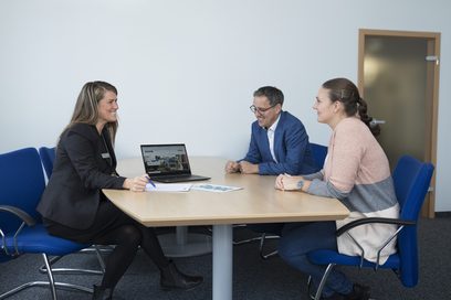 Eine Beraterin sitzt mit zwei Kunden am Tisch und zeigt ihnen ihre Investitionsmöglichkeiten auf einem Laptop und einem Flyer. (Foto: Axel Jusseit)