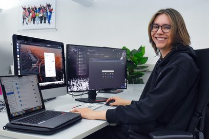 Ein junger Mann sitzt an einem Schreibtisch vor zwei Bildschirmen sowie einem aufgeklappten Laptop und bearbeitet digital einen Kundenauftrag.