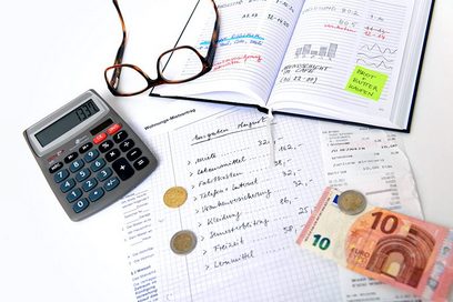 Das Foto zeigt einen Taschenrechnet, Kleingeld, einen Planner und einen Zettel über Finanzen sowie eine Brille.