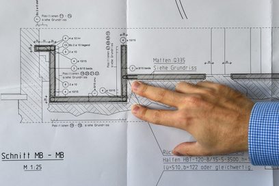 Das Bild zeigt den Grundriss eines Gebäudes aus der Frontalperspektive. Darauf liegt eine Hand, die drei Finger abspreizt. Am Handgelenk kann man erkennen, dass die Person ein Hemd trägt.