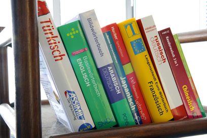 Mehrere Wörterbücher in den Fremdsprachen spanisch, chinesisch, russisch, französisch, italienisch, japanisch, arabisch, englisch und türkisch stehen schräg nebeneinander auf einem Stuhl.