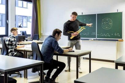 Eine Lehrkraft erklärt zwei jungen Männern im Klassenzimmer das Schalenmodell im Fach Chemie.