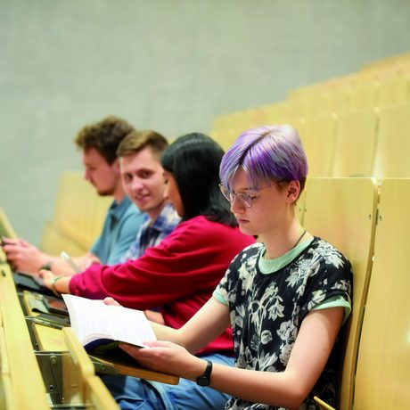 Vier junge Menschen sitzen in einem Hörsaal. Die vorderste Person blättert in einem Heft.