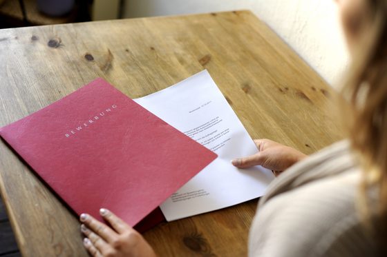 Man sieht einen Tisch und die Hände einer Person, die ein bedrucktes Blatt Papier in eine rote Mappe mit der Aufschrift "Bewerbung" steckt