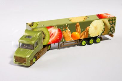 Ein grüner Spielzeug-Lastkraftwagen mit Auflieger auf einer weißen Fläche. Der Auflieger ist mit bunten Früchten bedruckt.