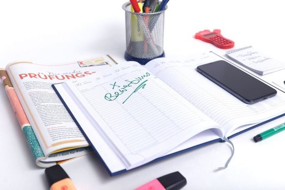 Ein Schreibtisch, auf dem eine Zeitschrift, ein aufgeschlagener Terminkalender, ein Smartphone, ein Stifteköcher und mehrere Textmarker liegen.