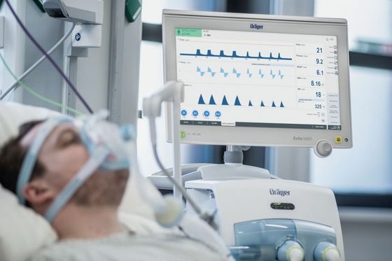 Zu sehen ist der Monitor des Beatmungsgerätes mit verschiedenen Messfunktionen. Im Vordergrund liegt ein Patient mit Atemmaske.