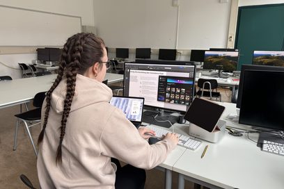 Rückansicht einer jungen Frau am Arbeitsplatz, die vor mehreren Bildschirmen sitzt und arbeitet. 