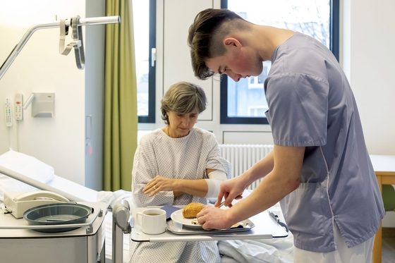 Ein junger Pfleger steht am Bett einer Patientin und bereitet ihr Frühstück vor.