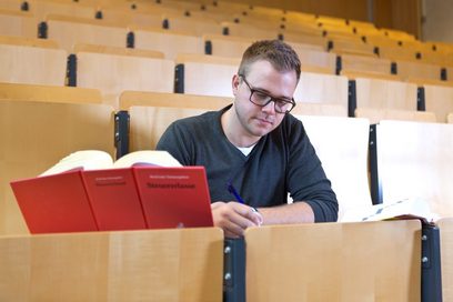 Ein junger Mann sitzt mit einem Gesetzbuch und Lernunterlagen in einem Hörsaal. (Foto: Axel Jusseit)