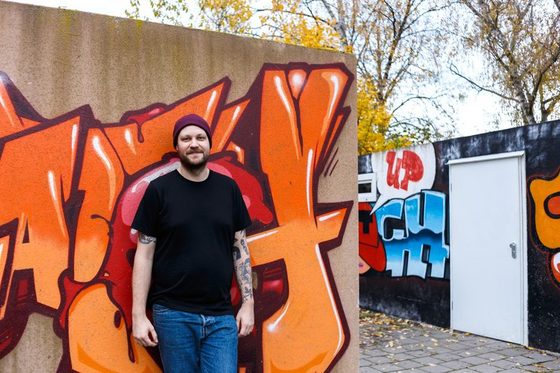 Daniel Norman steht vor einer Wand, die mit Grafitti besprüht ist.