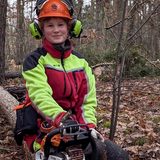 Die angehende Forstwirtin Rosali Ratai sitzt auf einem gefällten Baum, in voller Schutzkleidung und mit einer Motorsäge in der Hand.