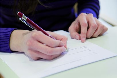 Jemand sitzt an einem Tisch mit einem Stift in der Hand und schreibt etwas auf ein Blatt Papier