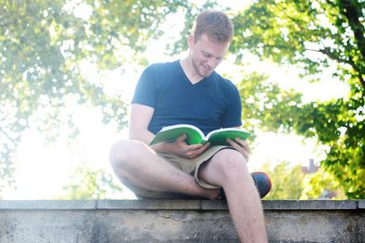 Junger Mann mit T-Shirt und kurzer Hose sitzt auf einer Mauer und liest in einem Buch.