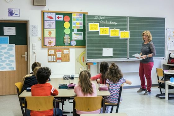 Eine Frau steht vor einer Tafel und schaut auf mehrere Kinder, die an einem Gruppentisch in einem Klassenraum sitzen.